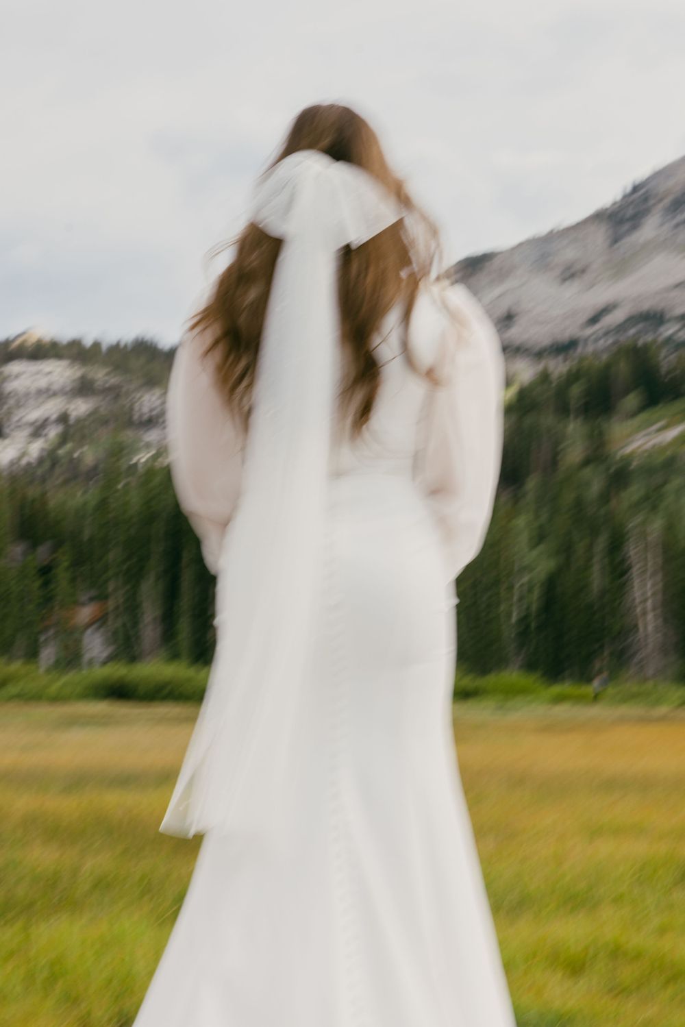 Motion-blurred image of bride in white dress standing in mountain meadow.