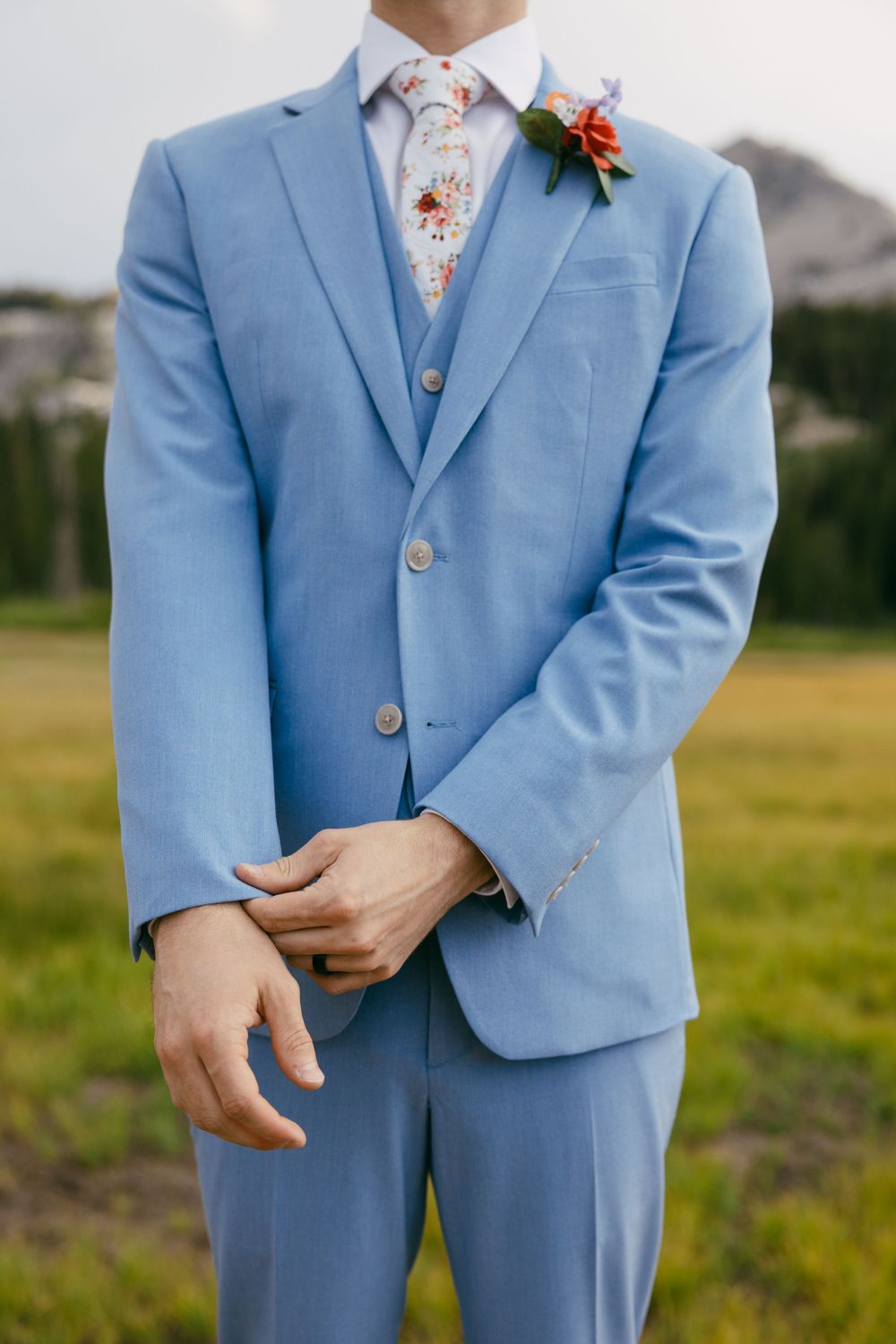 A light blue wedding suit with a floral tie and red boutonniere photographed against an outdoor background.