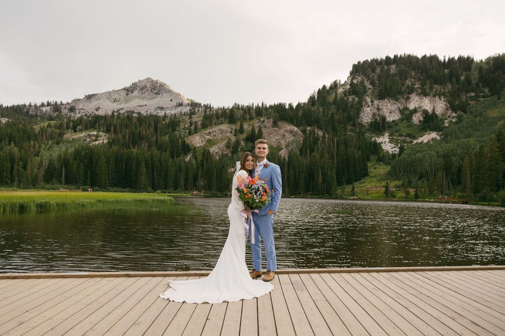 A wedding couple stands on a dock overlooking a mountain lake surrounded by evergreen forest.