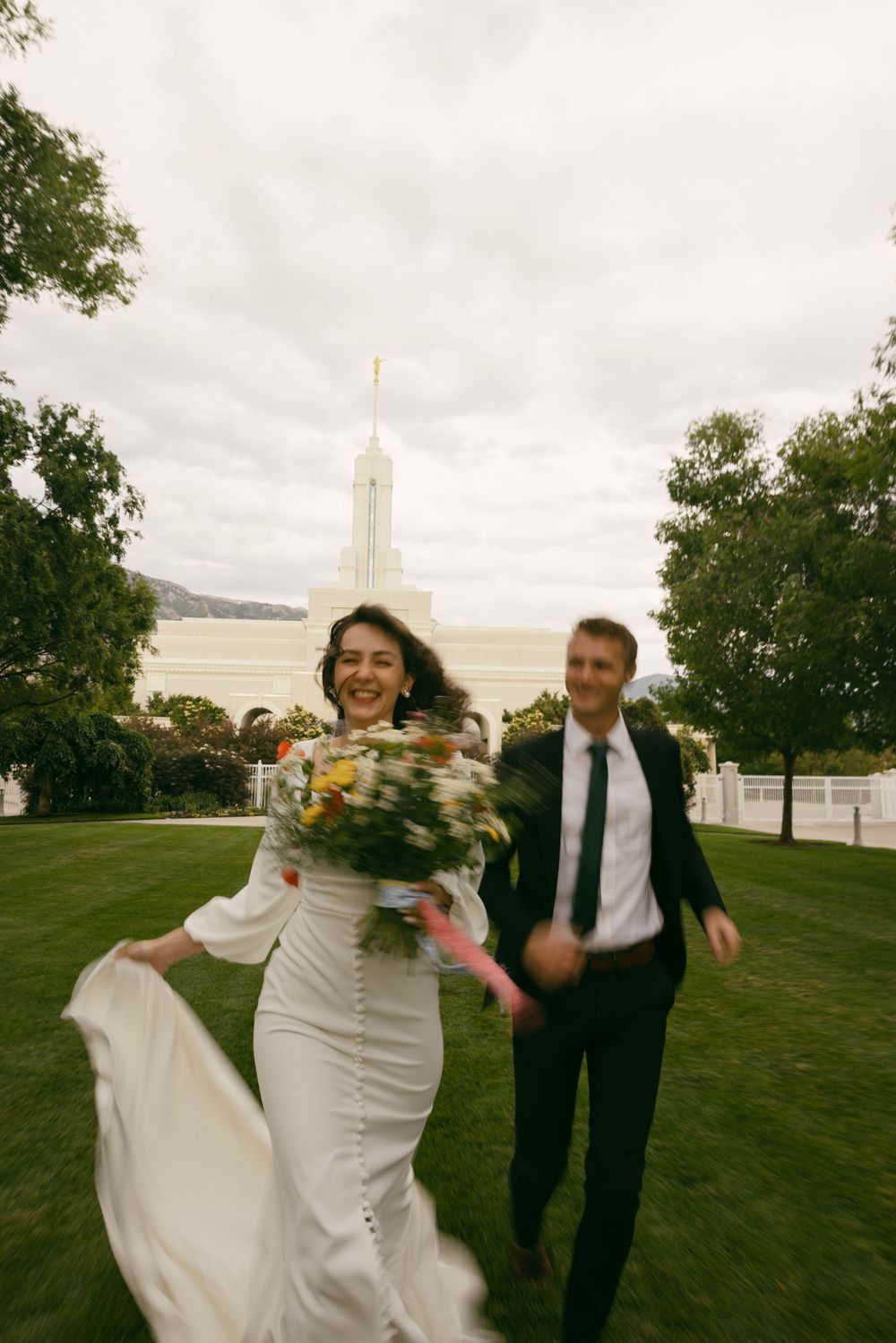 A joyful wedding couple runs across a lawn with a white temple building in the background.