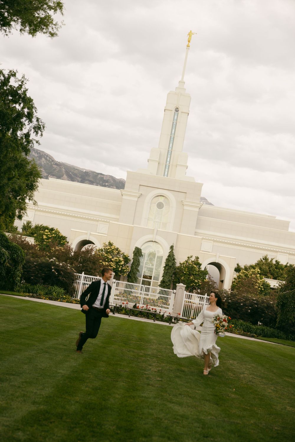 Wedding couple playfully jumping on grass with Mormon temple in background.