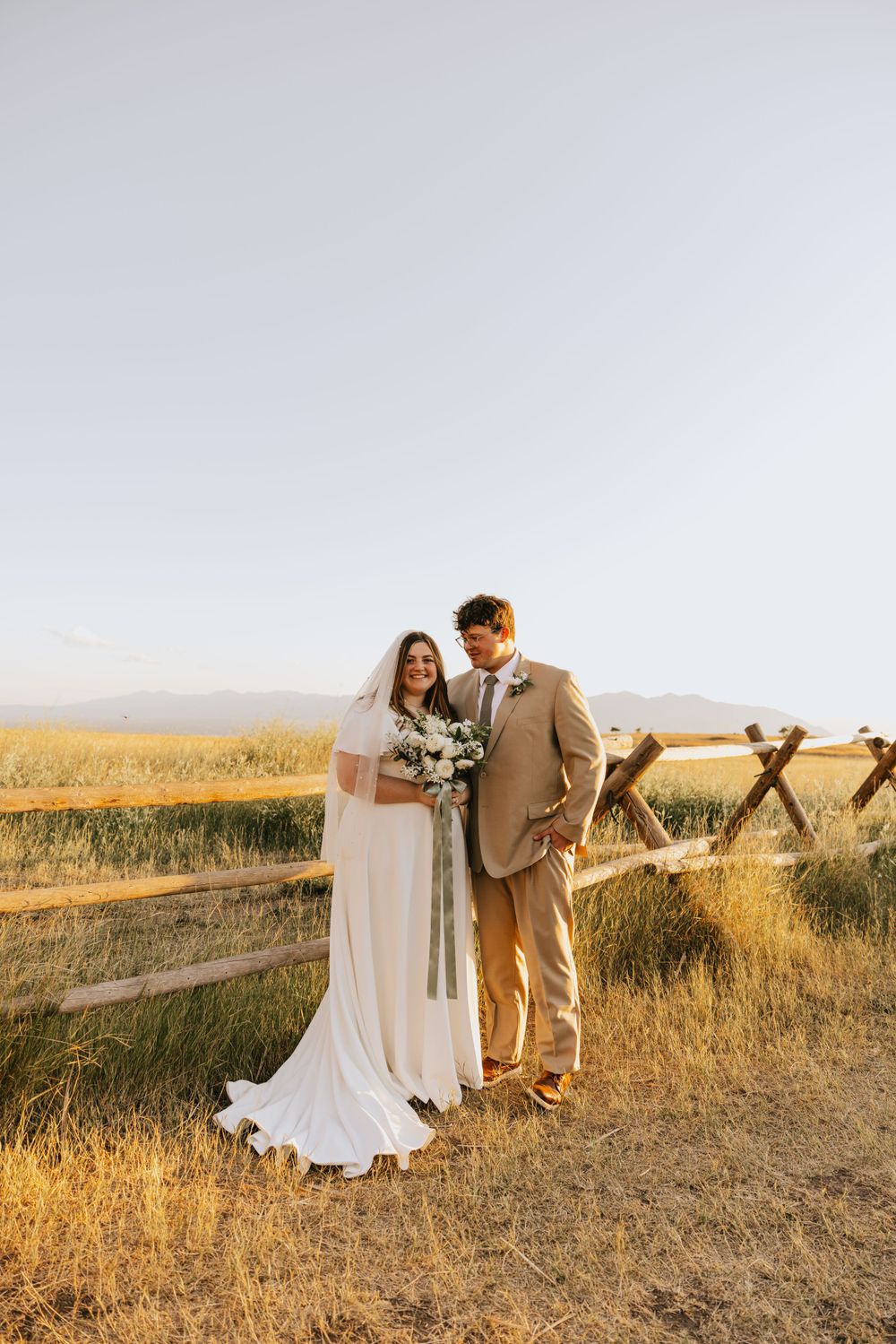 A sunset wedding portrait of a couple standing by a rustic wooden fence in a golden field.