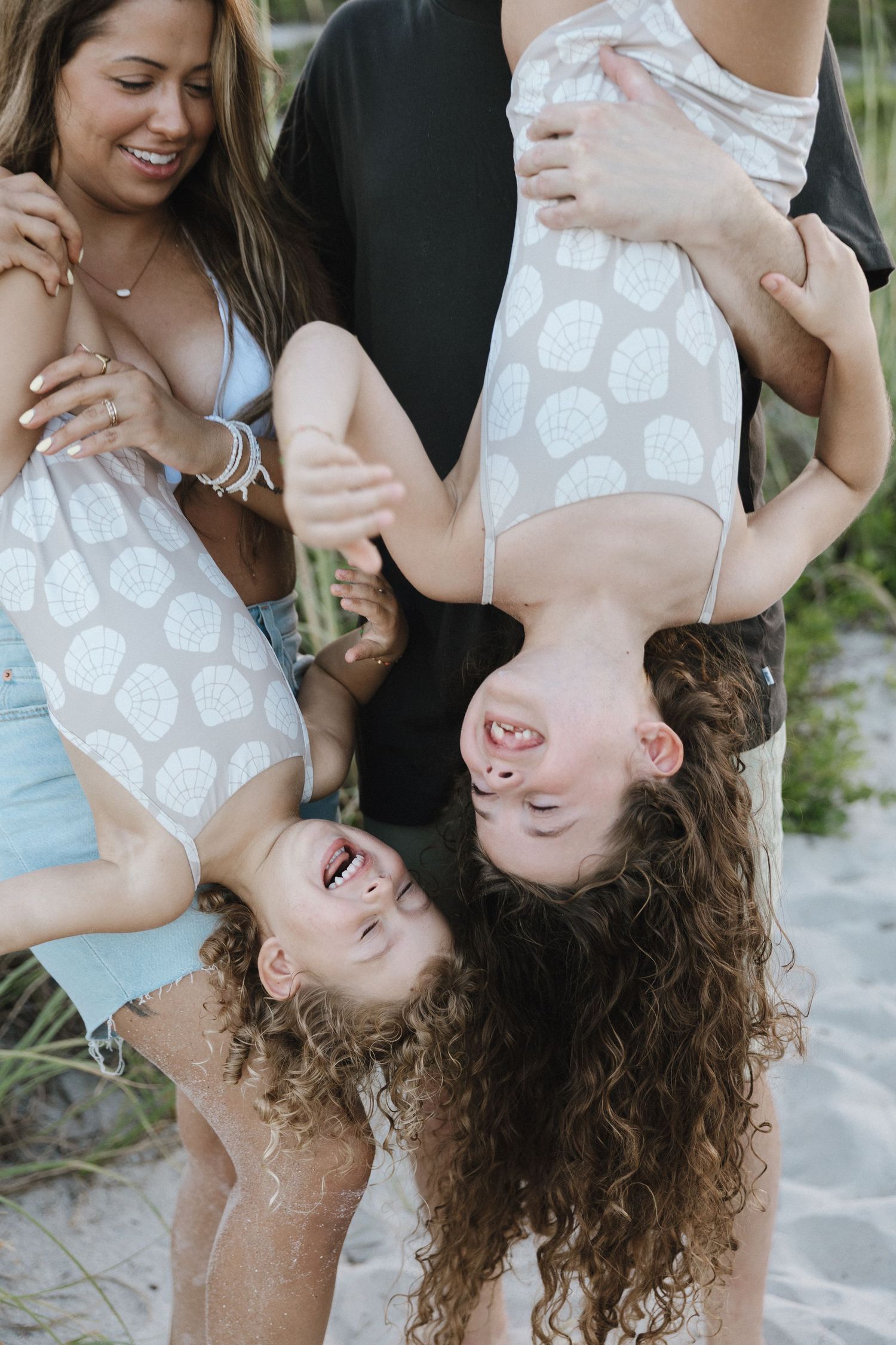 Parents playfully holding two laughing girls upside down on sandy beach near sea grass.