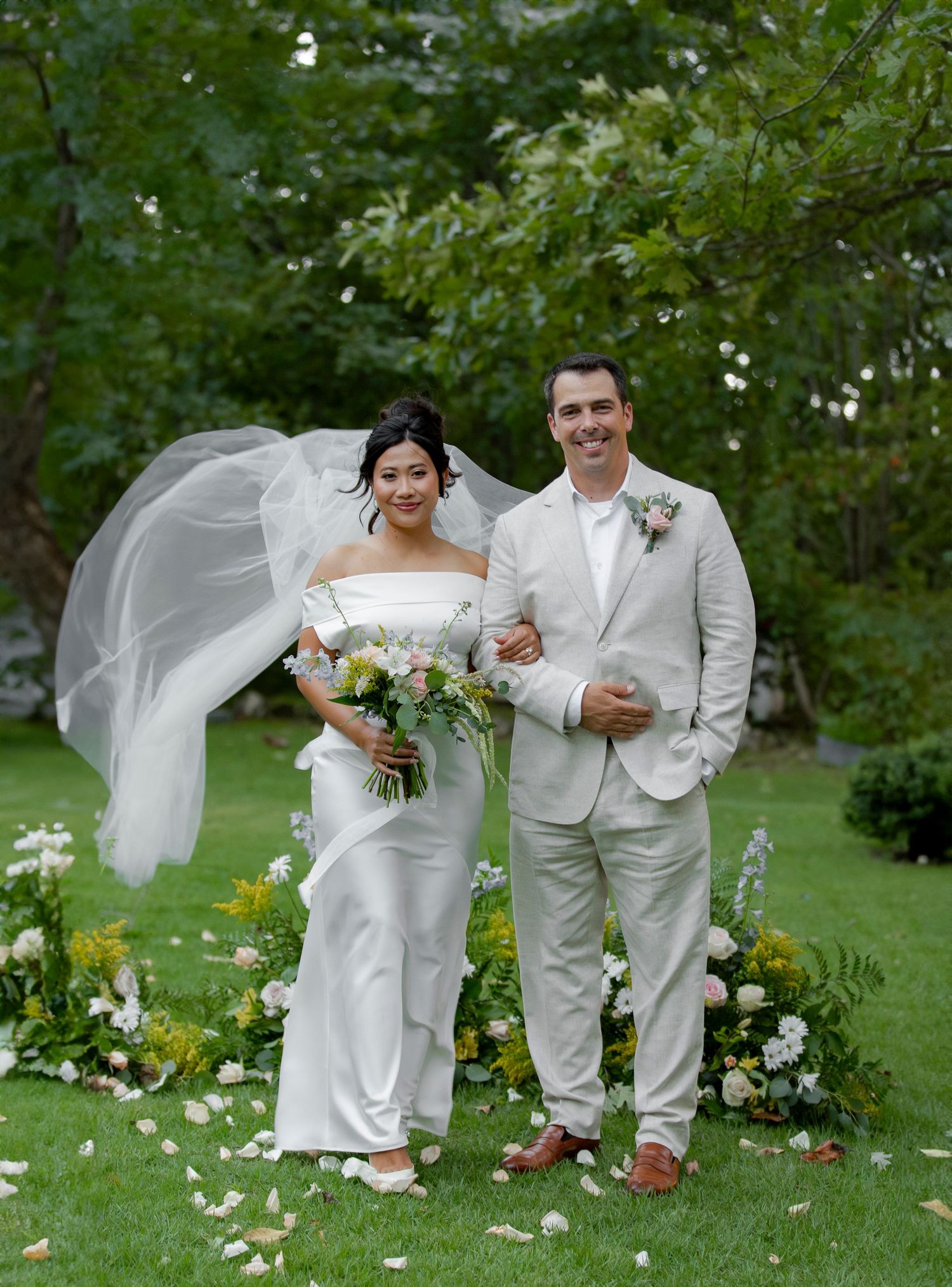A wedding couple poses in a garden setting with flowing white dress and light gray suit amid blooming flowers and greenery.