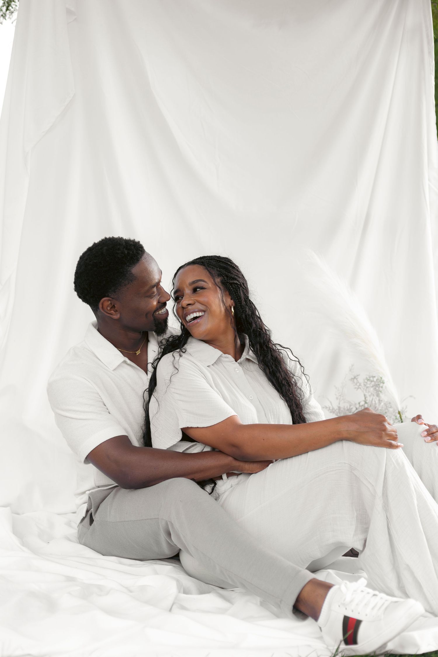 A loving couple dressed in white sit together and share a joyful moment against a light backdrop.