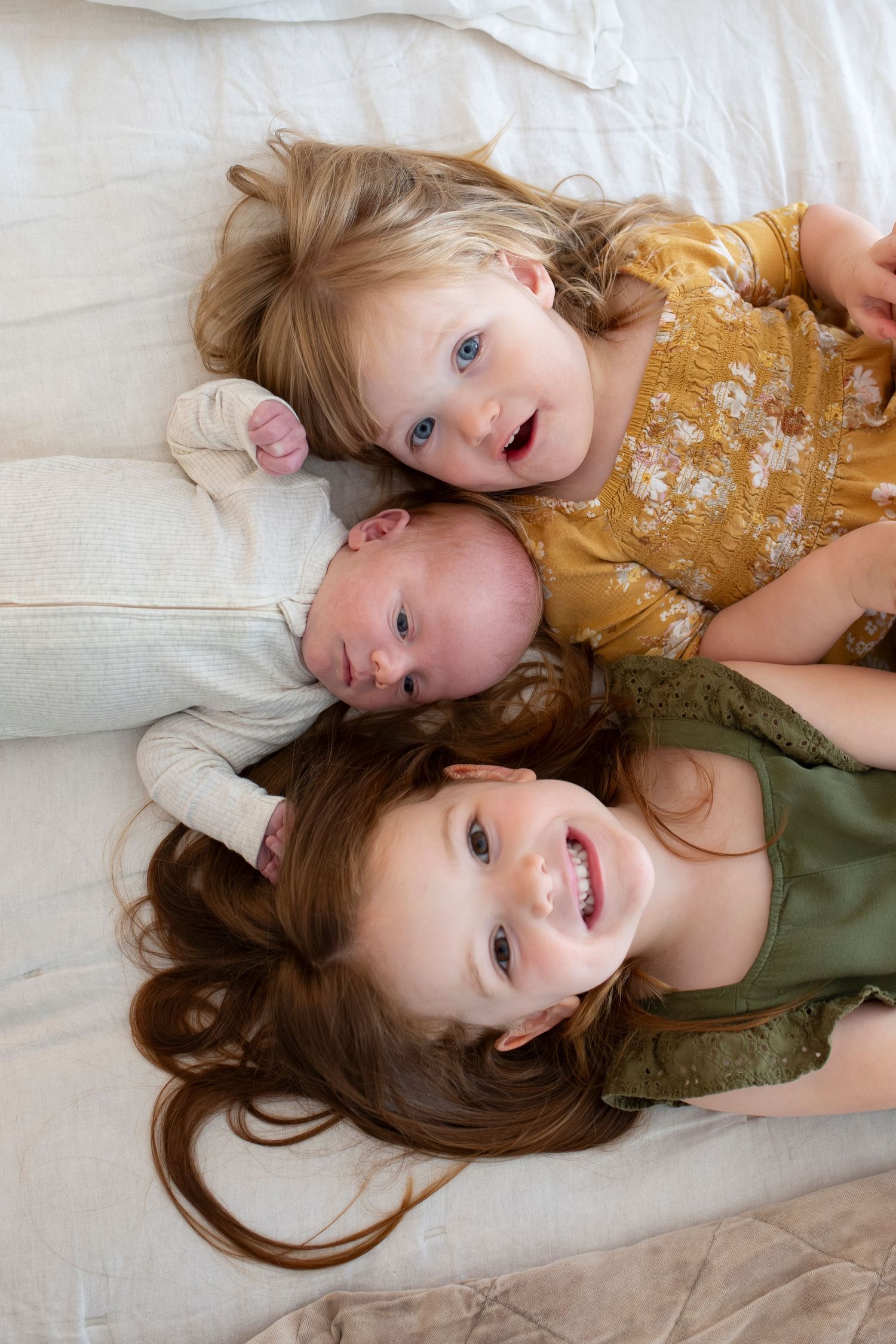 Two young girls and a newborn baby lying together on a white bed, smiling in a sweet sibling portrait.