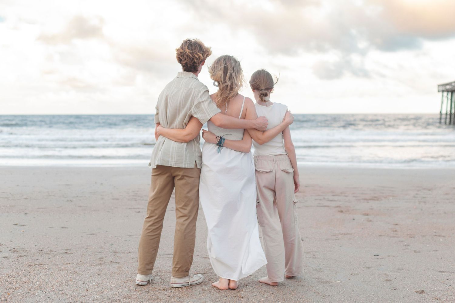 Three people embrace while watching the sunset on a peaceful beach with cloudy skies and ocean waves.