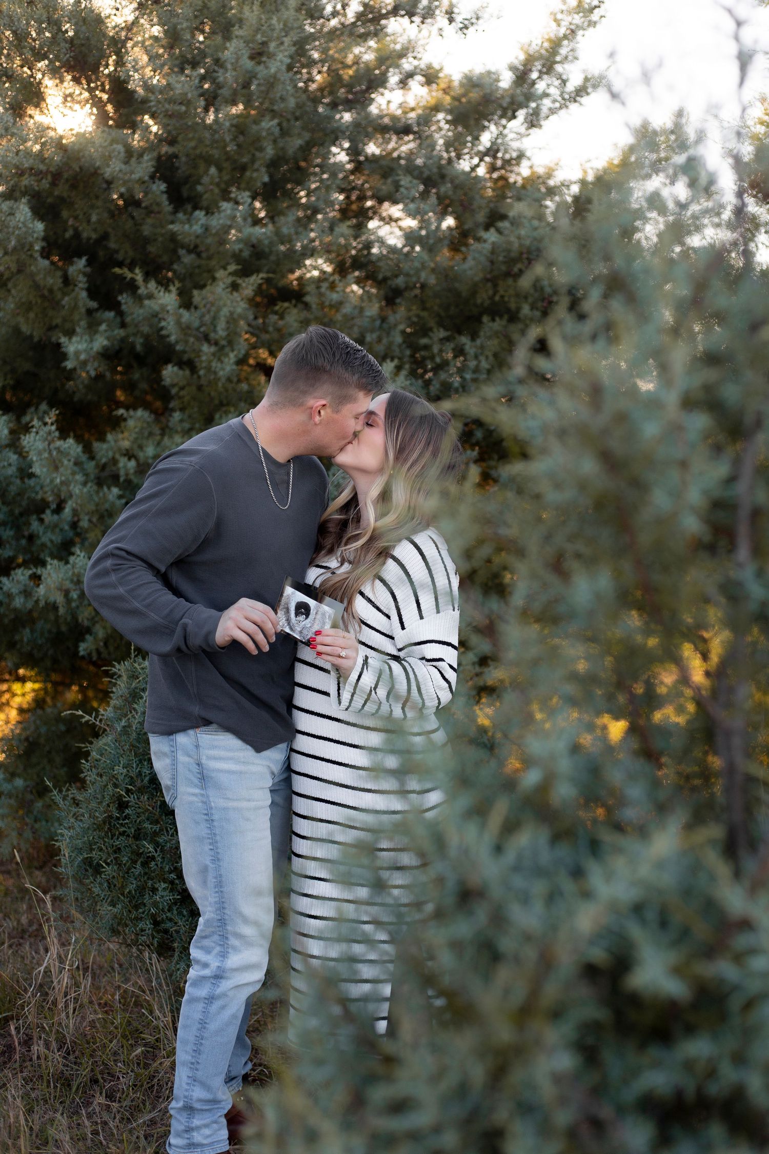 A couple shares a romantic moment while holding ultrasound photos among evergreen trees at sunset.