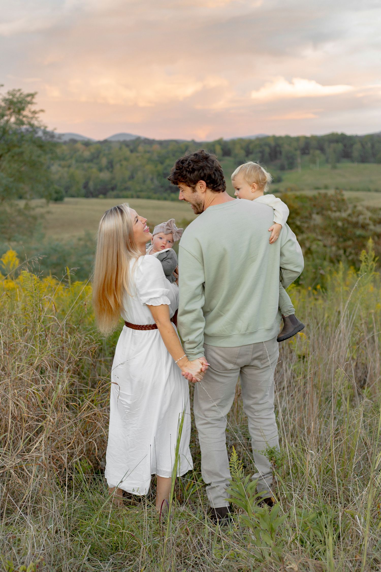 A family stands together in a field at sunset with rolling hills and yellow wildflowers in the background.