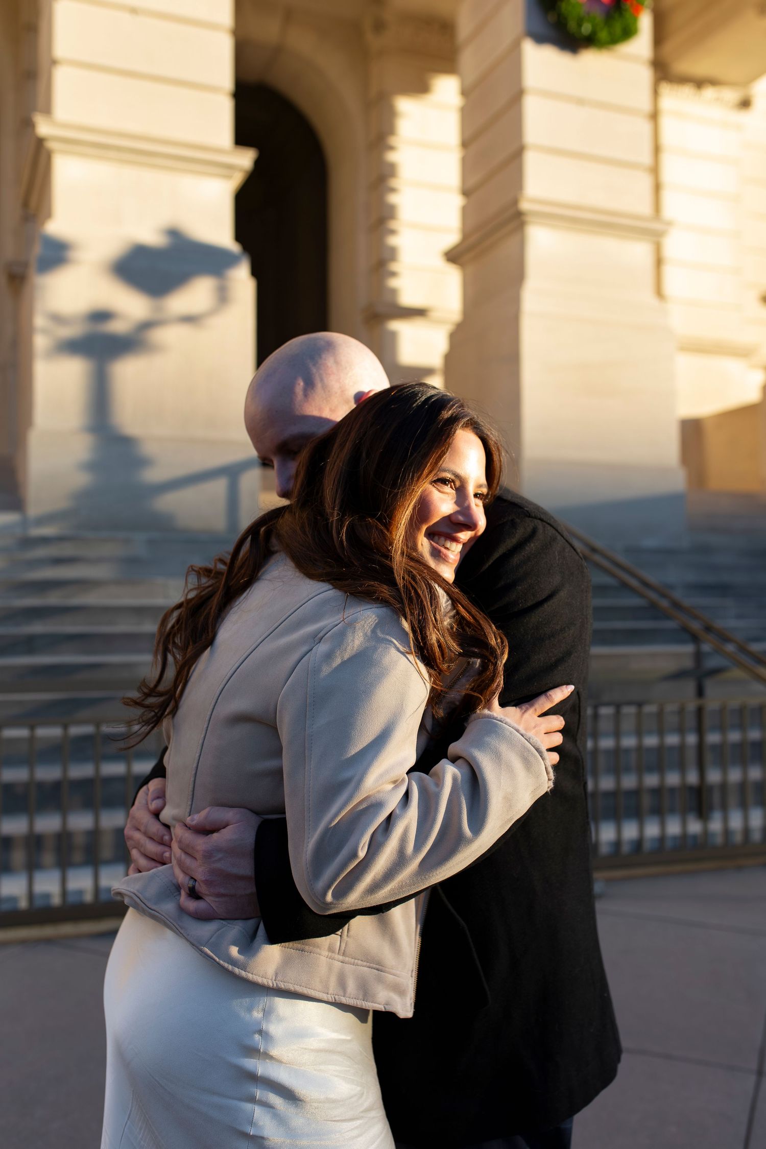 A smiling woman in a white jacket embraces a bald man in a black suit outside a classical stone building.