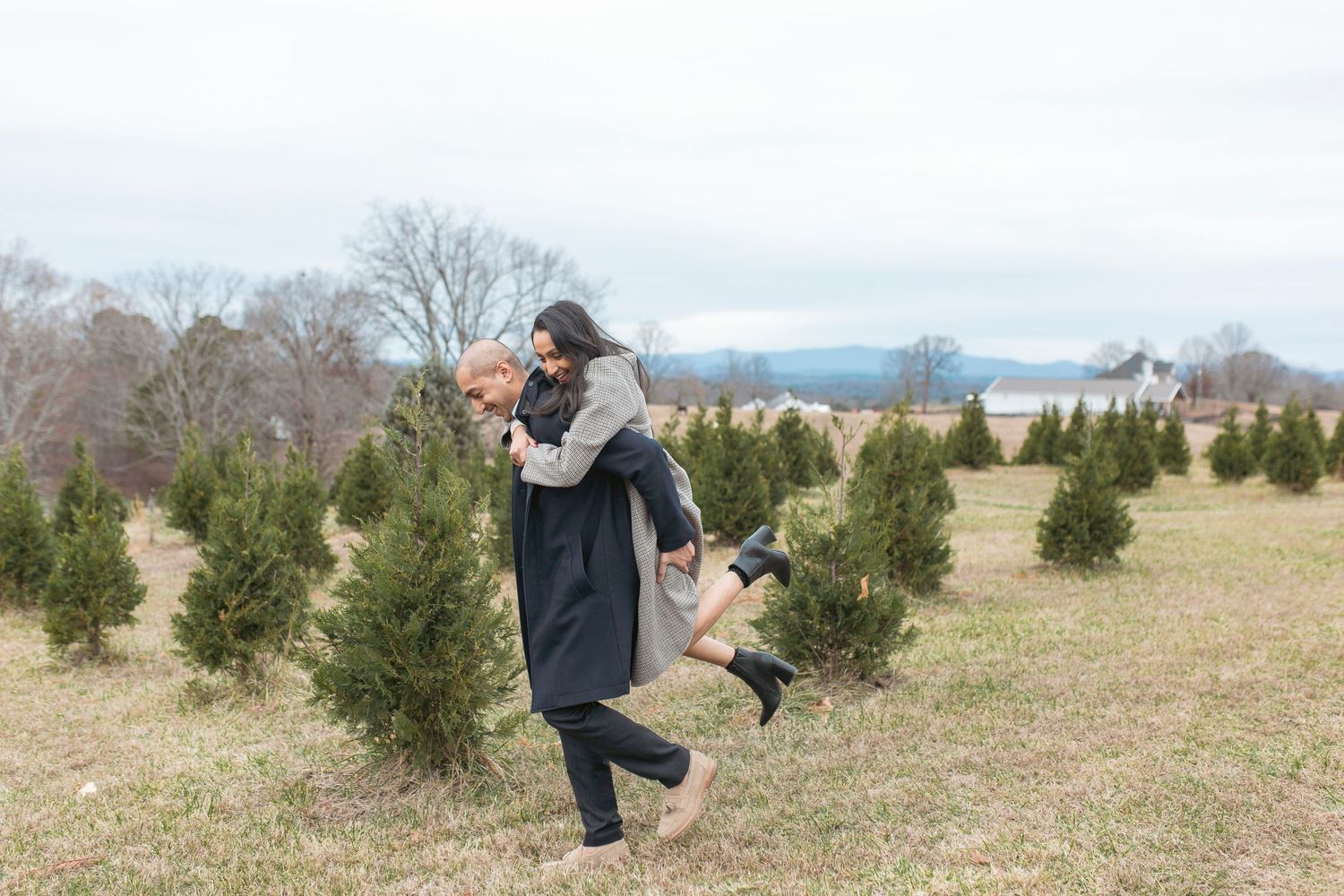 Two people embrace among evergreen trees at a Christmas tree farm on a cloudy winter day.