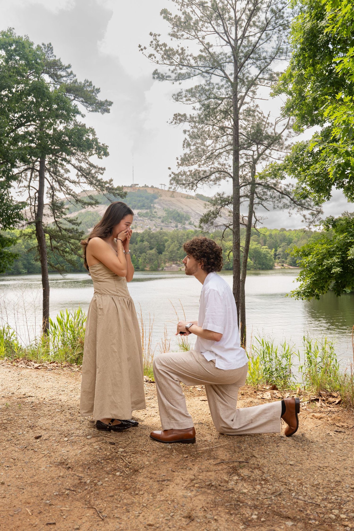 Man proposes on one knee to emotional woman by a serene lake surrounded by lush green trees.