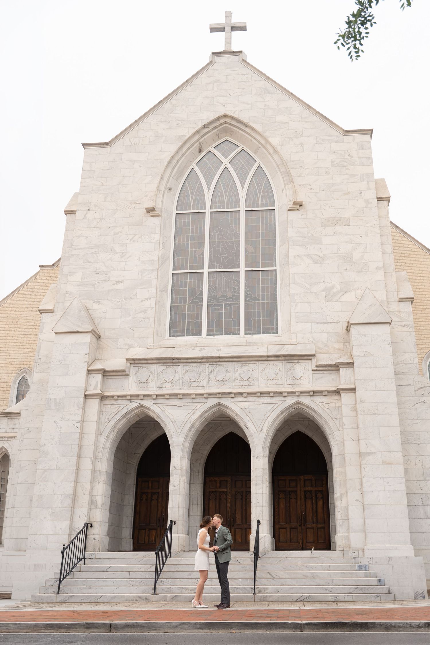 Couple shares a romantic moment on the steps of a grand white stone church with Gothic arched windows and a cross.