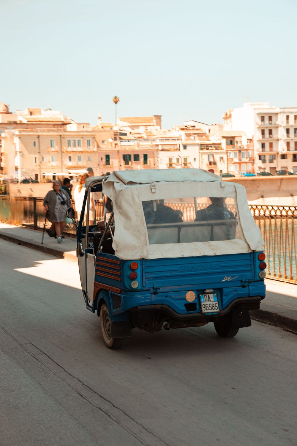 Blue vintage pickup truck parked along a waterfront promenade with buildings in the background.