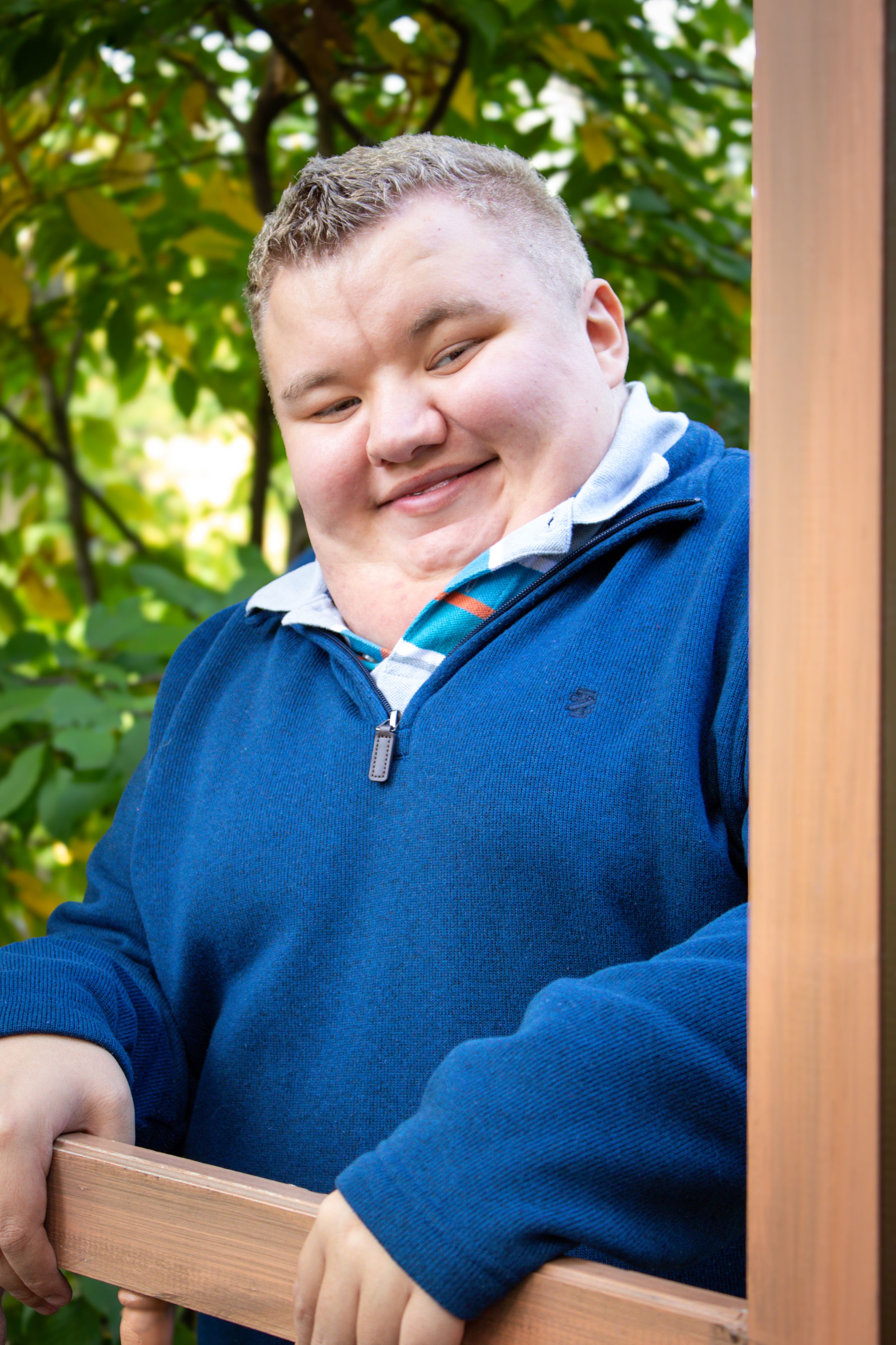 Senior portrait of a boy peeking playfully around the corner of a door outside surrounded by green trees.