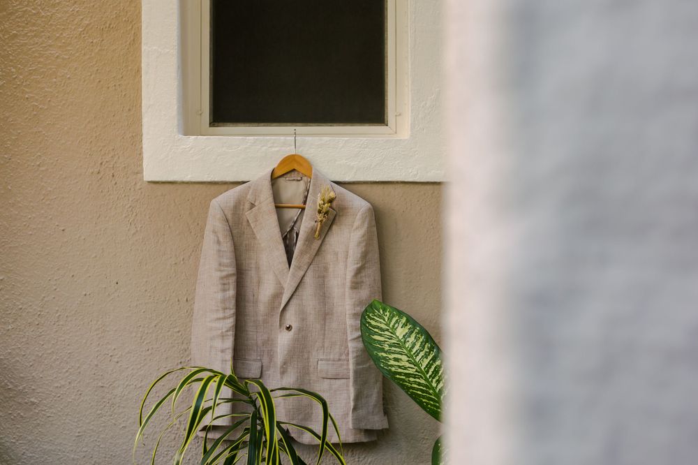 A beige suit jacket hanging outside near a white window frame with tropical leaves.