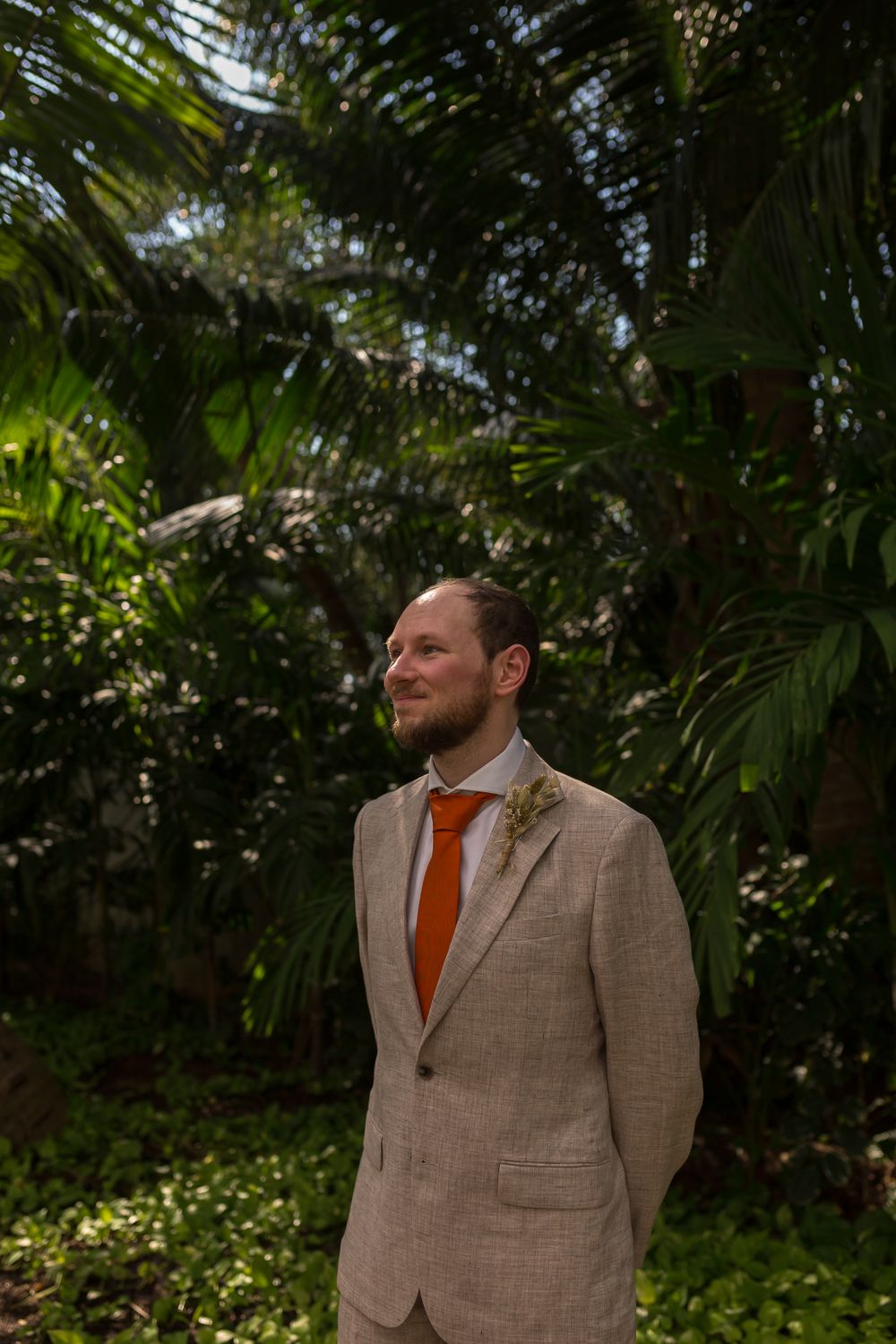 A groom in a beige suit with orange tie stands thoughtfully in a lush tropical garden setting.