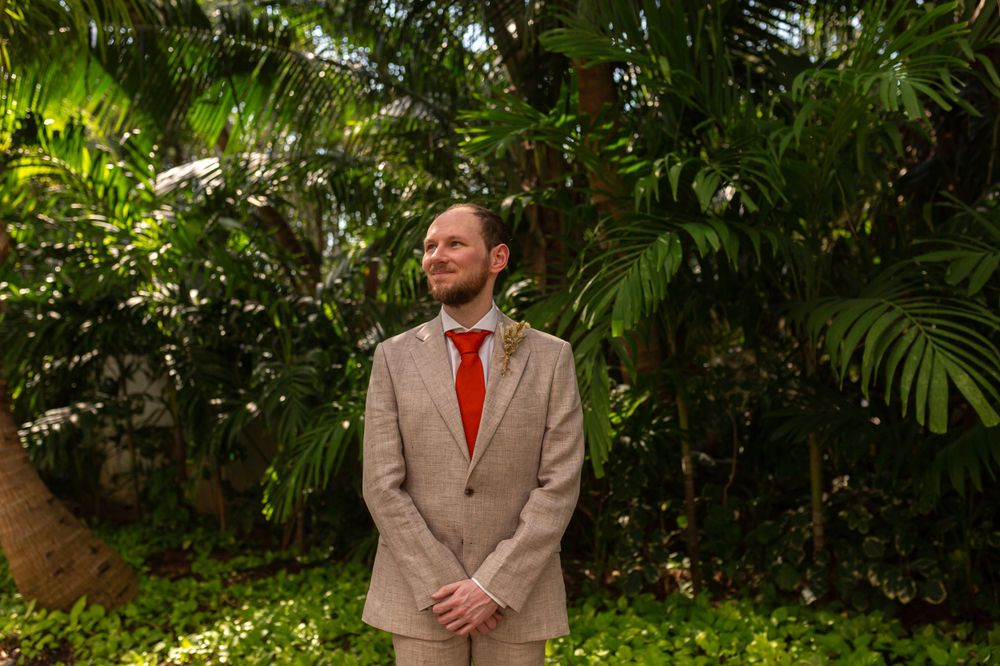 Wedding attendee in tan suit and red tie poses against verdant tropical foliage backdrop.
