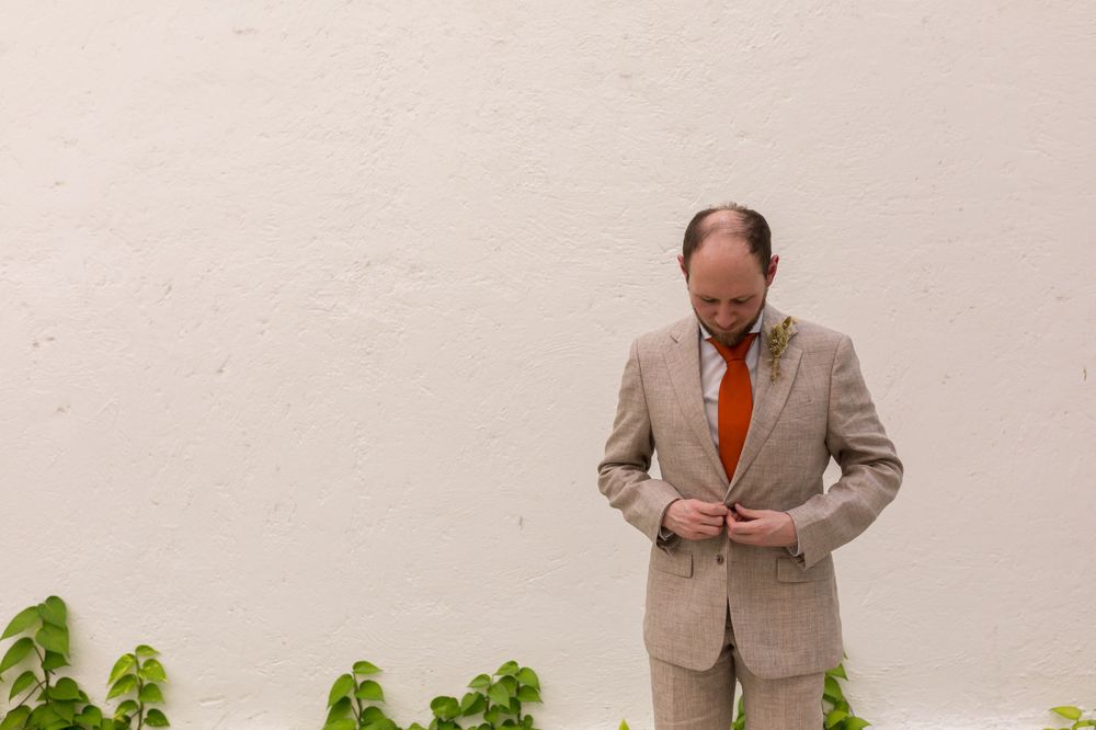 Person in beige suit adjusts jacket buttons against white wall with greenery below.