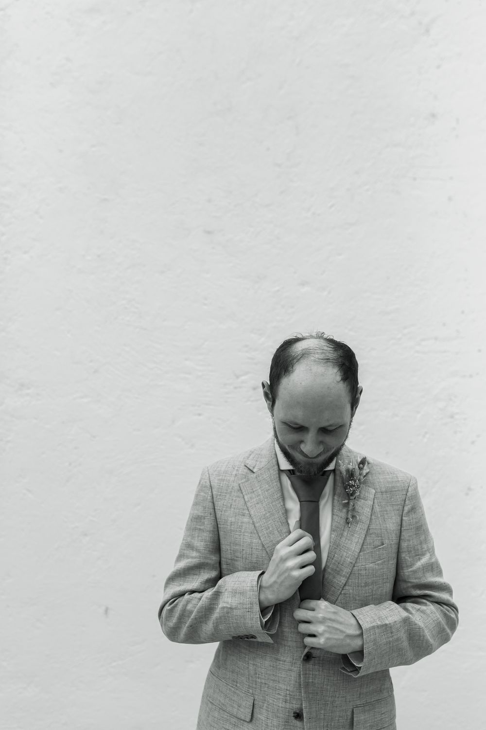 Black and white portrait of person in light suit adjusting tie against white wall.