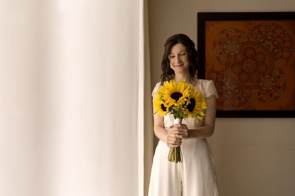 Bride in white dress holds vibrant sunflower bouquet near window with wall art.