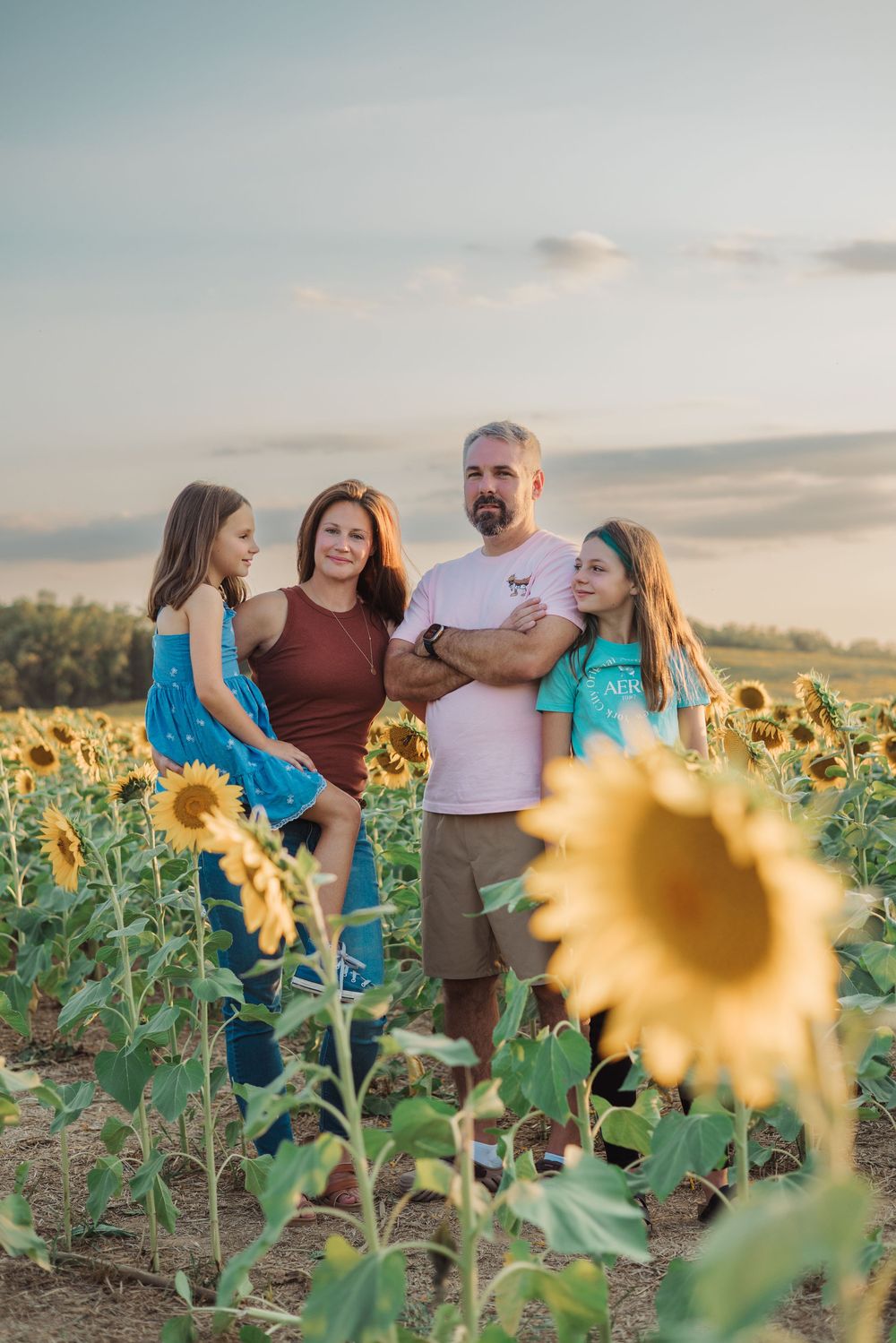 A family poses together in a vibrant sunflower field during golden hour sunset with soft clouds in the background.