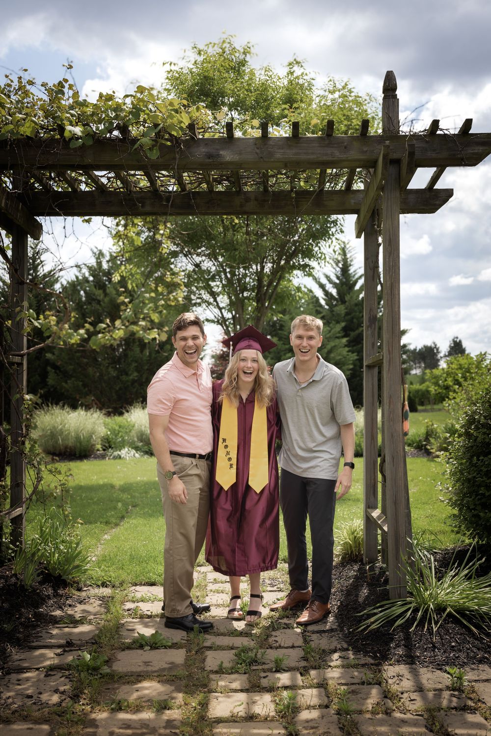 Graduation photo under a wooden garden pergola with climbing vines and plants on a cloudy day.