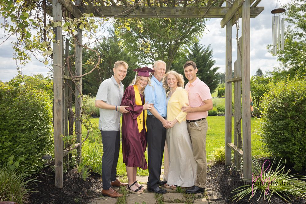 A group stands together under a wooden garden arbor surrounded by lush greenery on a sunny day.