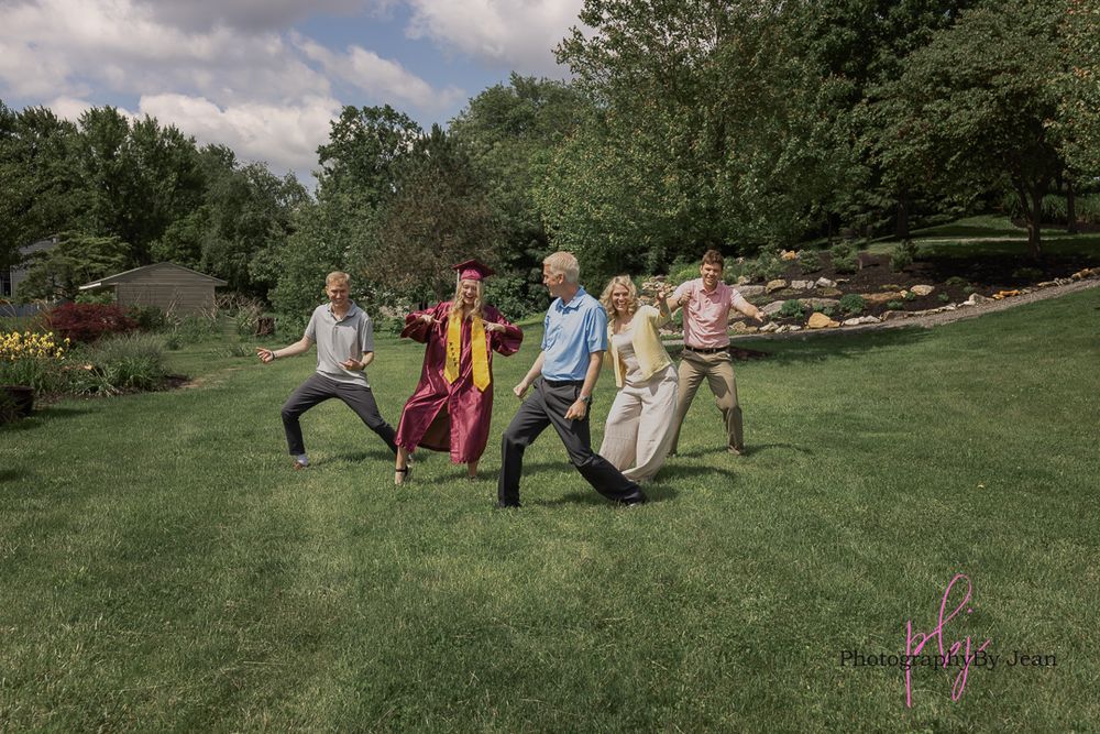A group of people strike warrior yoga poses while practicing outdoors on a green lawn surrounded by trees.