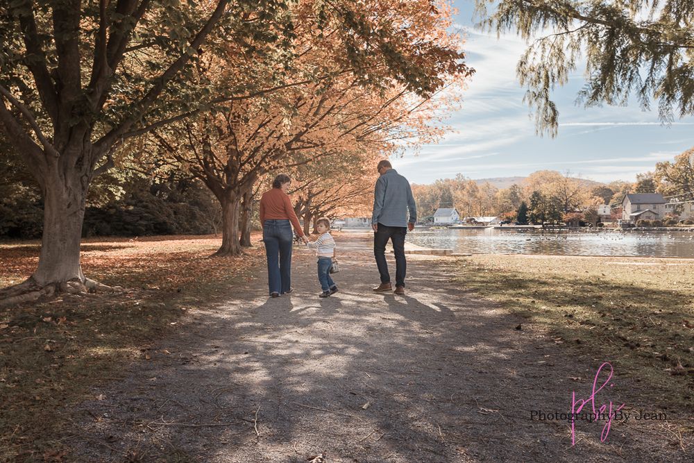 A family walks together on a tree-lined path by a lake during autumn with colorful fall foliage.