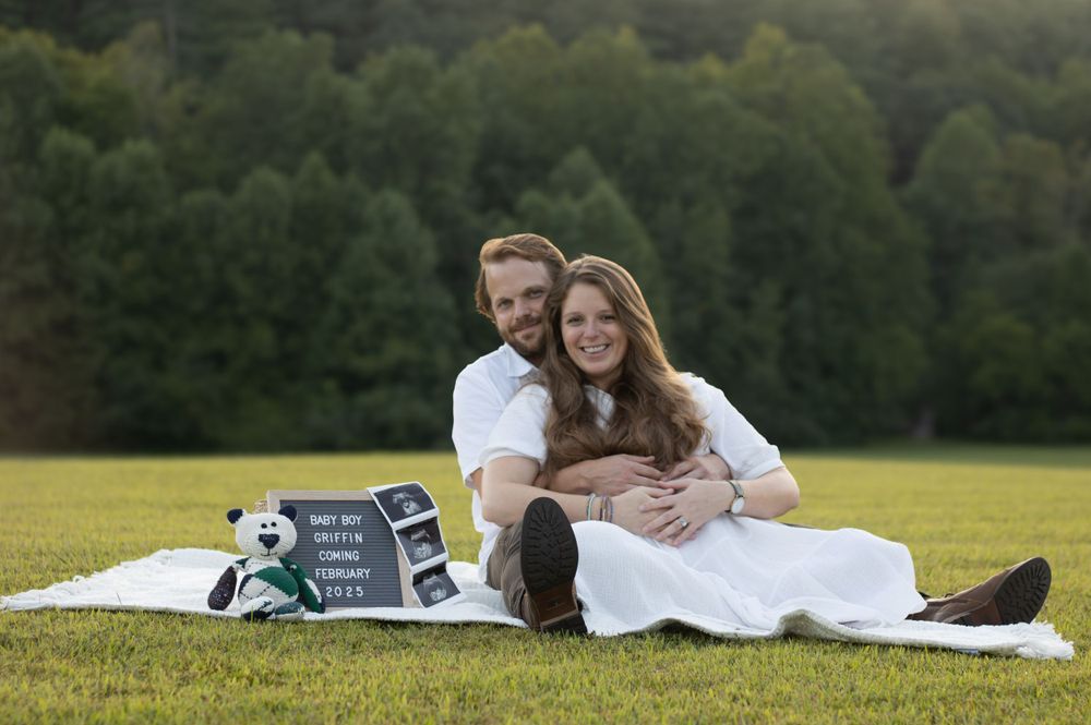 Couple lying on blanket in grassy field with trees in background.
