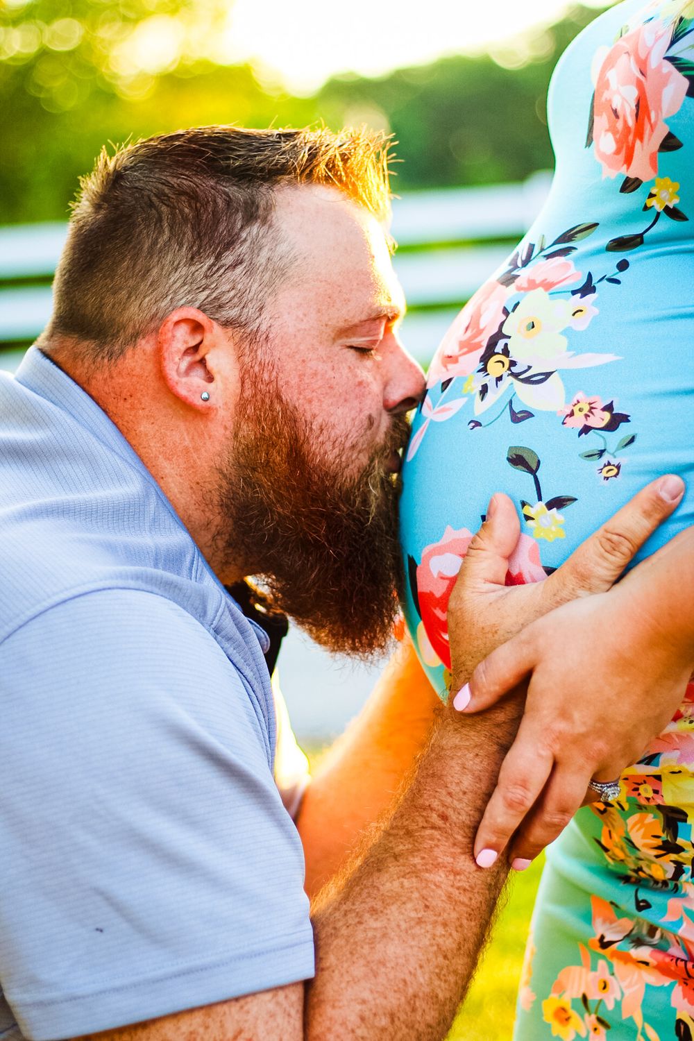 Candid Maternity photo of a man holding and kissing his wife's pregnant belly with the sunlight shining on a fence in the background.