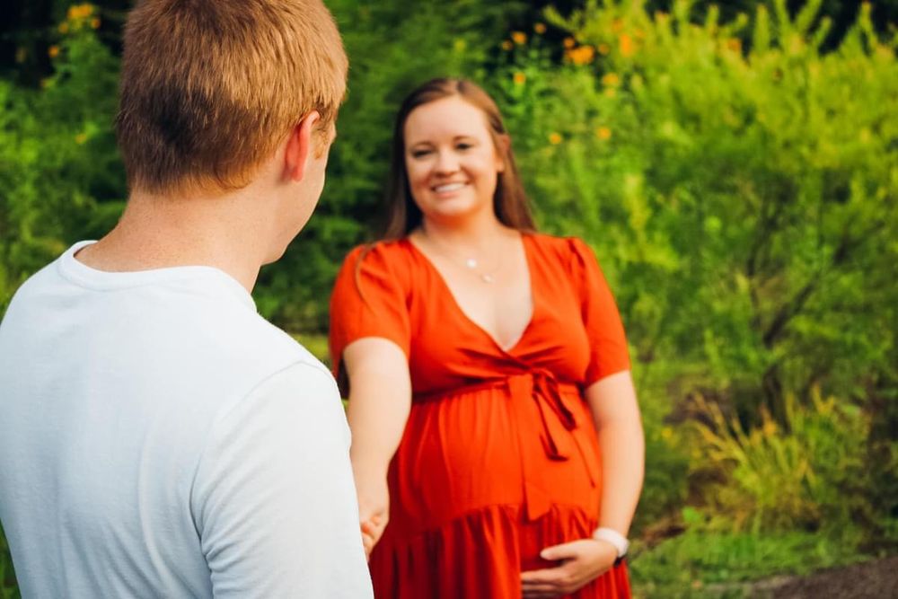 A maternity candid of a couple holding hands and smiling at each other with greens in the background.