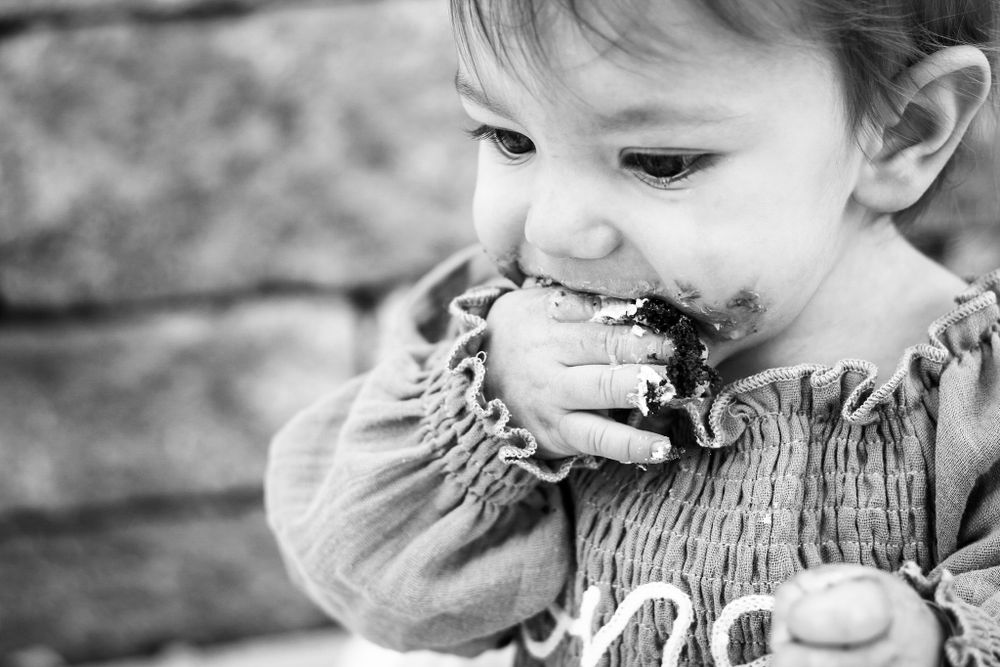 A black and white lifestyle image of a baby's One-year cake smash photoshoot as she shoves cake into her mouth.