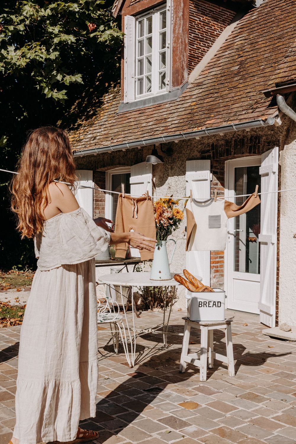 Woman in white dress hanging laundry on clothesline outside rustic brick cottage with bread box and flowers on table.