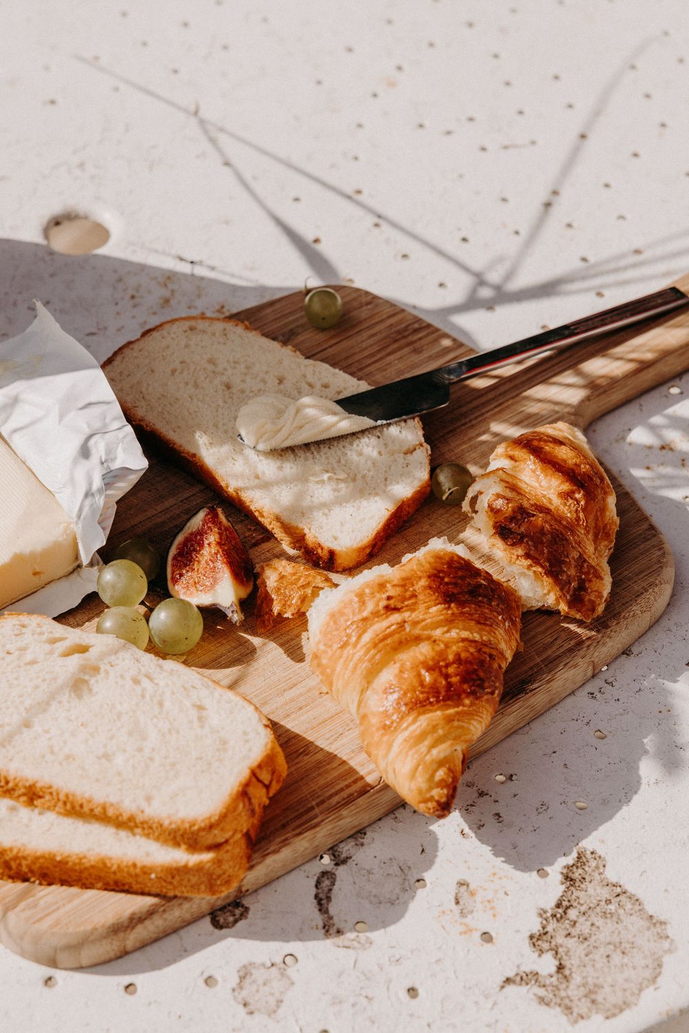 Wooden cutting board with croissant, sliced bread, brie cheese, figs, and green grapes in bright sunlight.