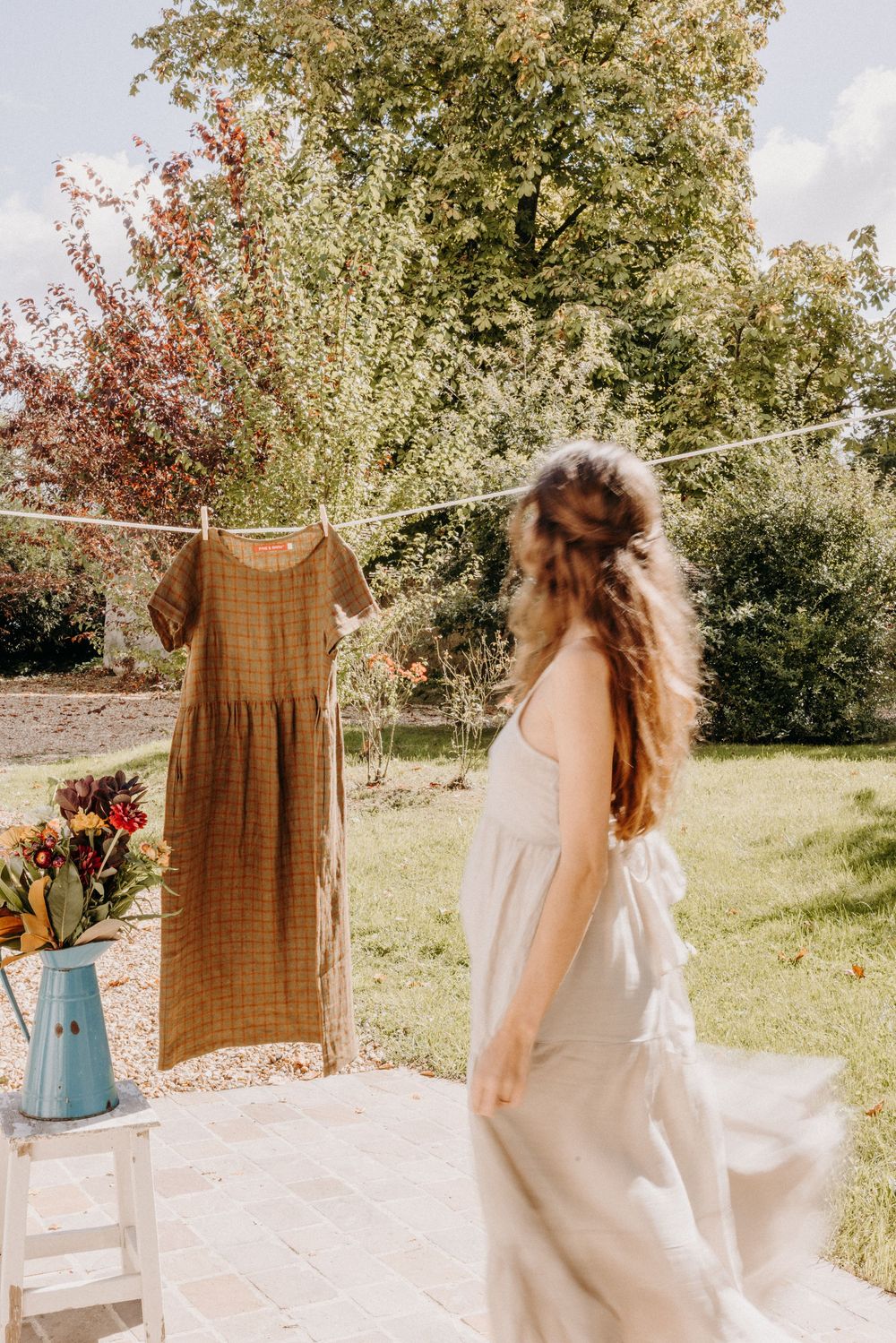 Woman in white dress stands in sunny garden beside clothesline with brown linen dress hanging near blue vase of flowers.