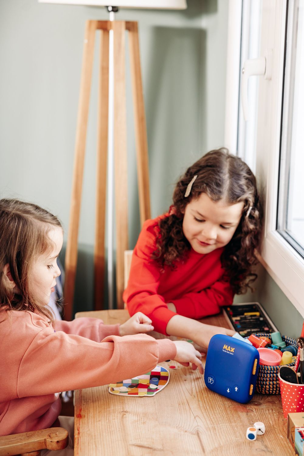 Two young girls playing with colorful educational toys and a blue MAX device at a wooden table by a window.