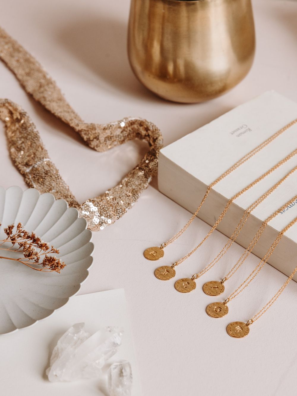 Gold coin necklaces displayed on white box with sequin ribbon, crystal, and golden vase on pink background.