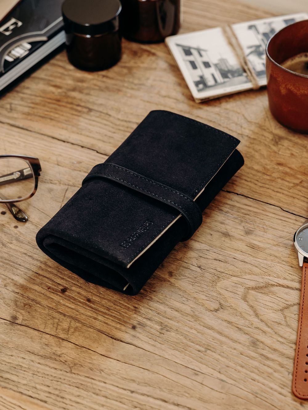 Black leather roll wallet on wooden desk with glasses, watch, coffee, and vintage photos.