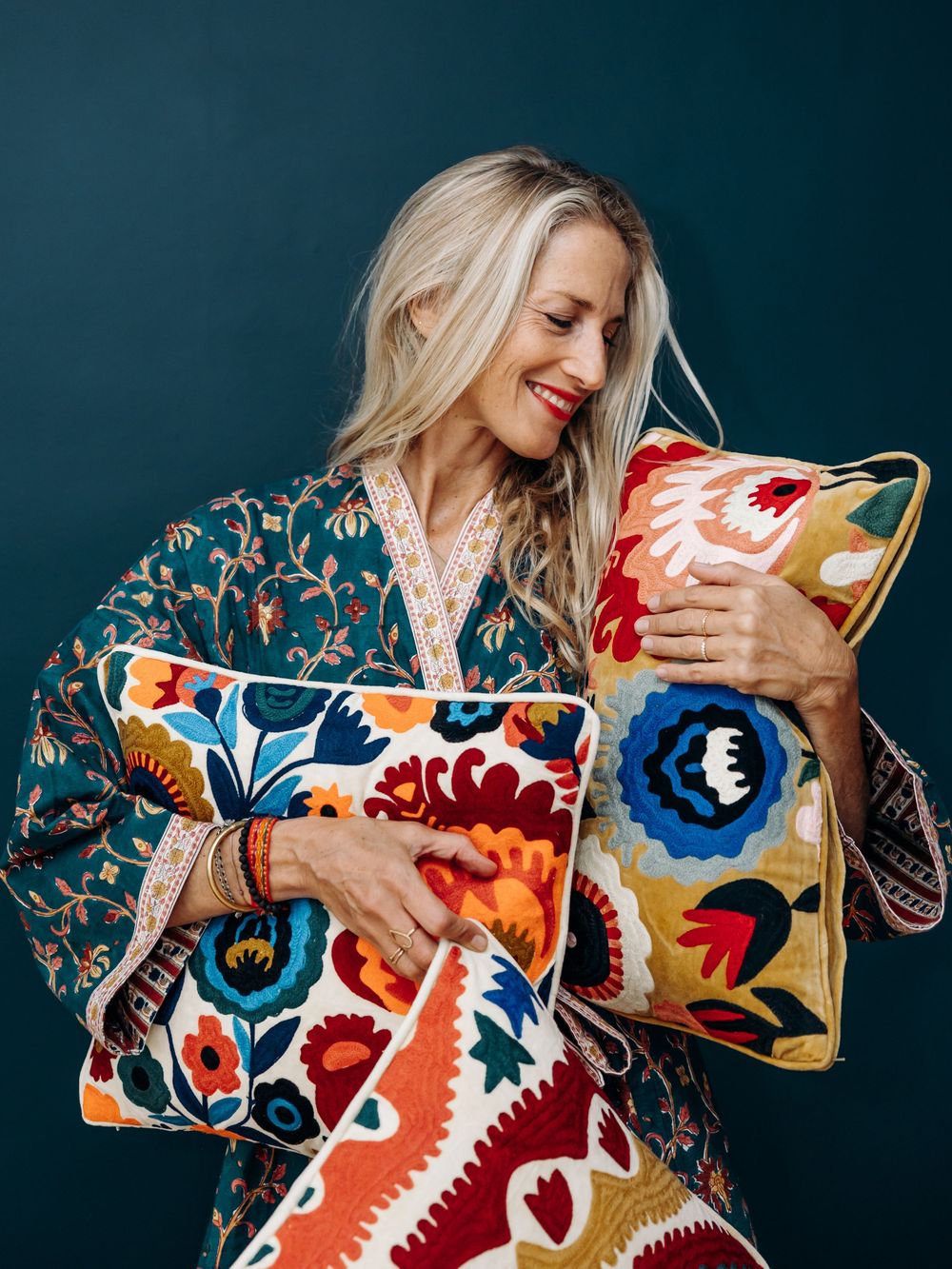 Smiling blonde woman in colorful floral kimono holding vibrant embroidered decorative pillows against teal background.
