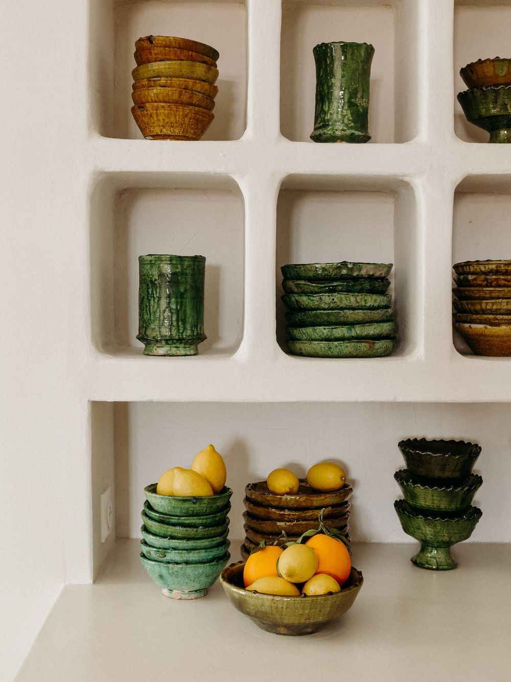 White shelves display stacked green and amber ceramic bowls, vases, and plates alongside fresh lemons and oranges in rustic pottery.