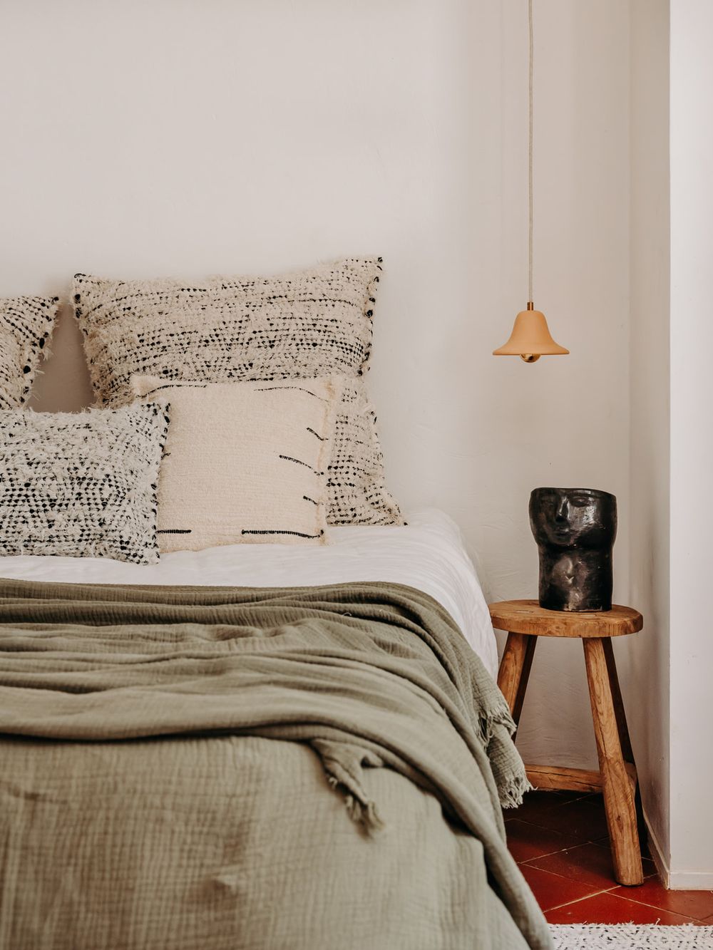 Cozy bedroom with sage green linen blanket, patterned pillows, rustic wood stool nightstand, and pendant light.