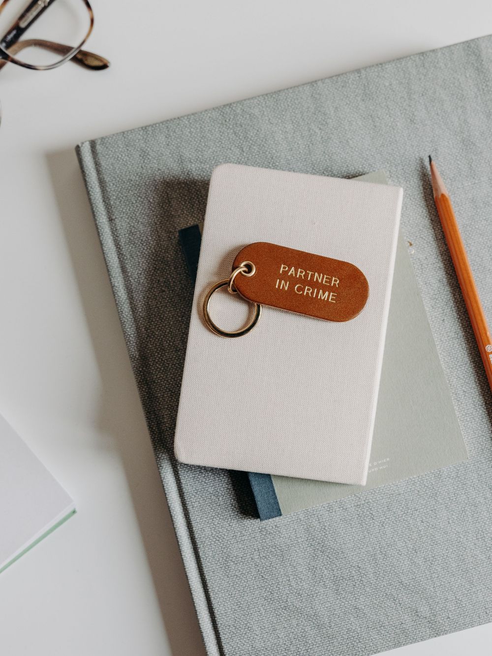 Leather keychain reading Partner In Crime resting on a small notebook with pencil and glasses on a white desk.