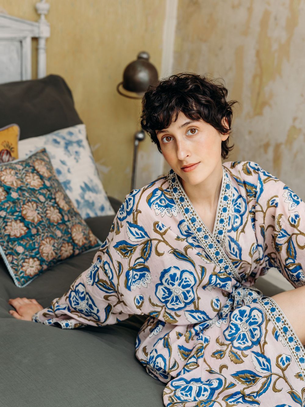 Person in floral blue and white wrap dress sitting on a gray sofa with decorative pillows in a vintage room.
