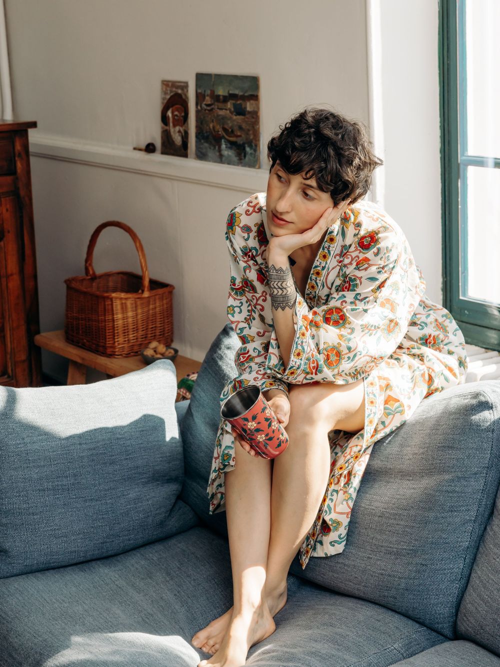 Woman in floral robe sits on gray sofa holding a mug, gazing thoughtfully in a bright, cozy living room.