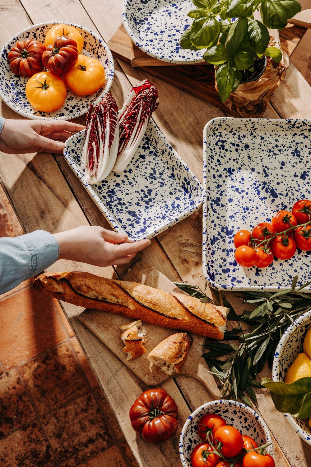 Hands arranging radicchio on speckled blue and white ceramic dishes with tomatoes, baguette, and fresh basil on a wooden table.