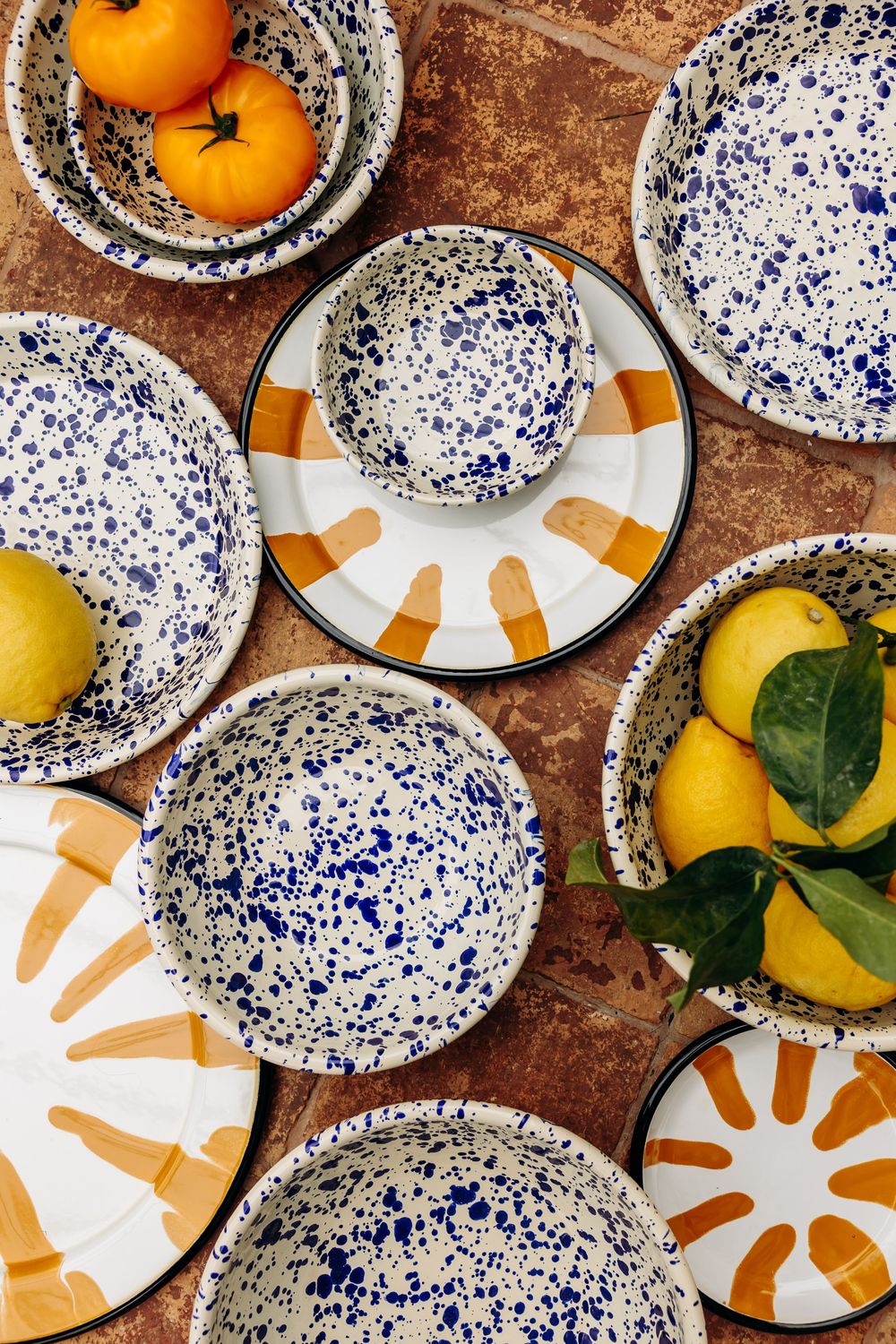Blue splatter ceramic plates and bowls with orange sun designs displayed with fresh lemons and oranges on rustic surface.