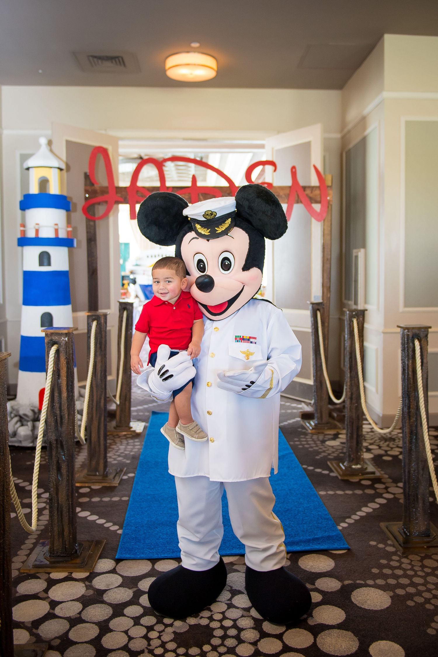 Mickey Mouse costumed character in cruise director attire stands next to a lighthouse prop in a restaurant setting.