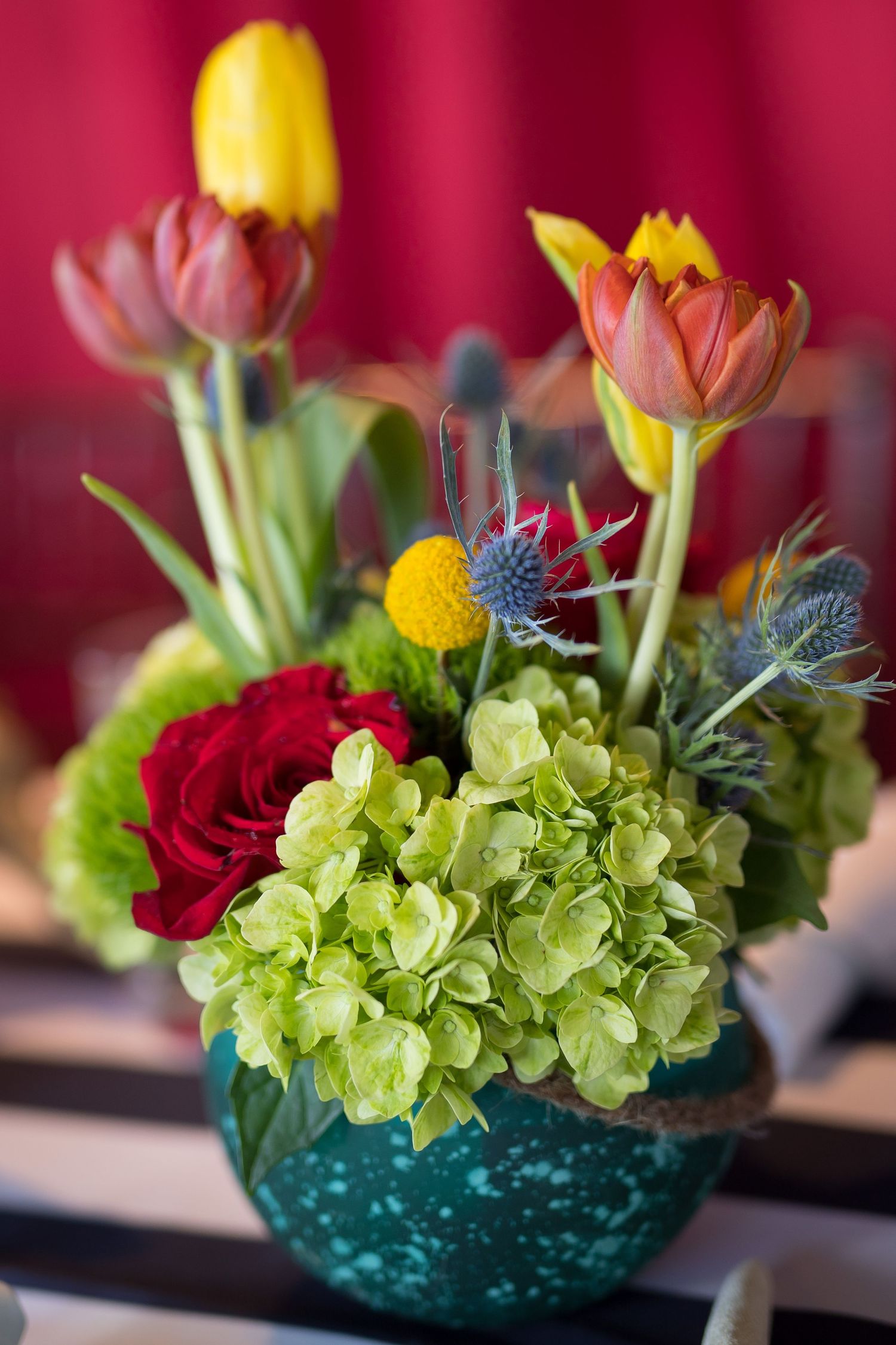 Colorful flower arrangement with red roses, green hydrangeas, yellow tulips and thistle in a teal glass vase against red backdrop.